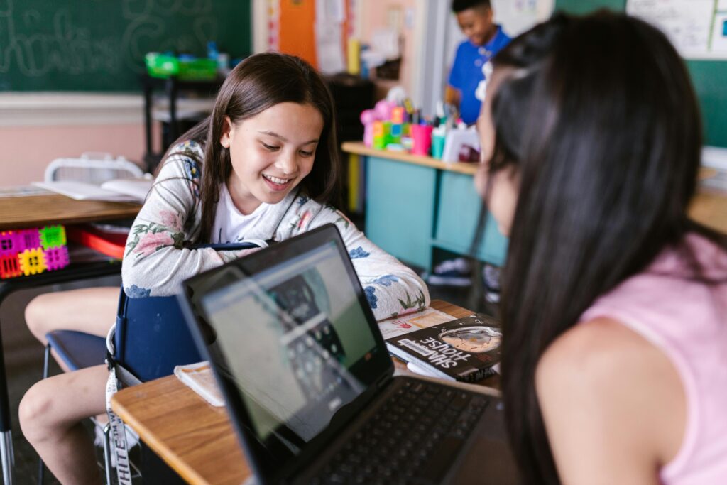 Duas alunas em sala de aula interagem com um notebook em cima da carteira escolar, enquanto sorriem e conversam. Ao fundo, o ambiente é decorado com materiais escolares e quadro-negro, representando um cenário de aprendizado colaborativo e digital.