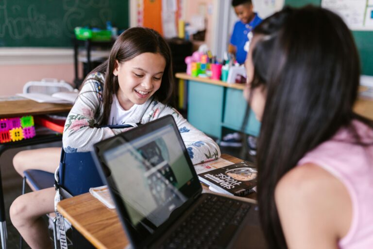 Duas alunas em sala de aula interagem com um notebook em cima da carteira escolar, enquanto sorriem e conversam. Ao fundo, o ambiente é decorado com materiais escolares e quadro-negro, representando um cenário de aprendizado colaborativo e digital.