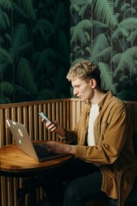 Homem jovem trabalhando remotamente em um café, utilizando um notebook e um smartphone ao mesmo tempo — representando multitarefa, mobilidade e conectividade no ambiente de trabalho digital.