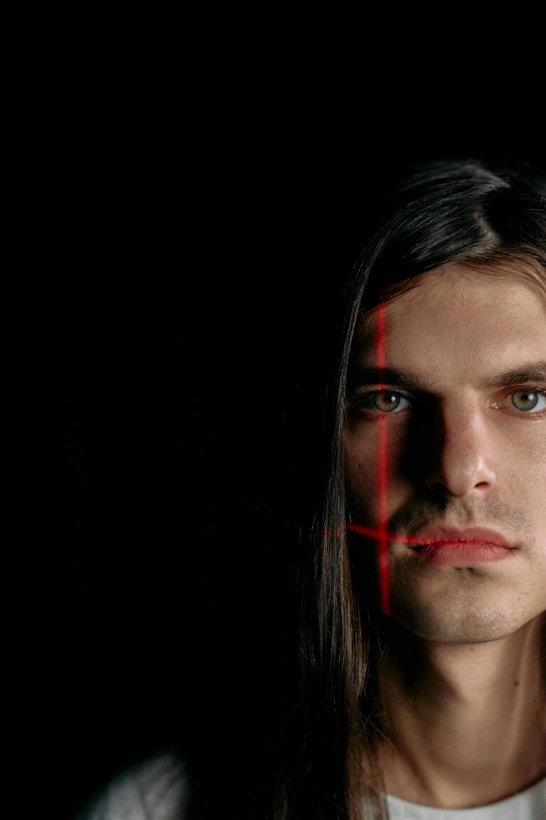 Rosto parcialmente iluminado por feixes de luz em forma de cruz, representando análise facial por biometria comportamental em um ambiente escuro e controlado.