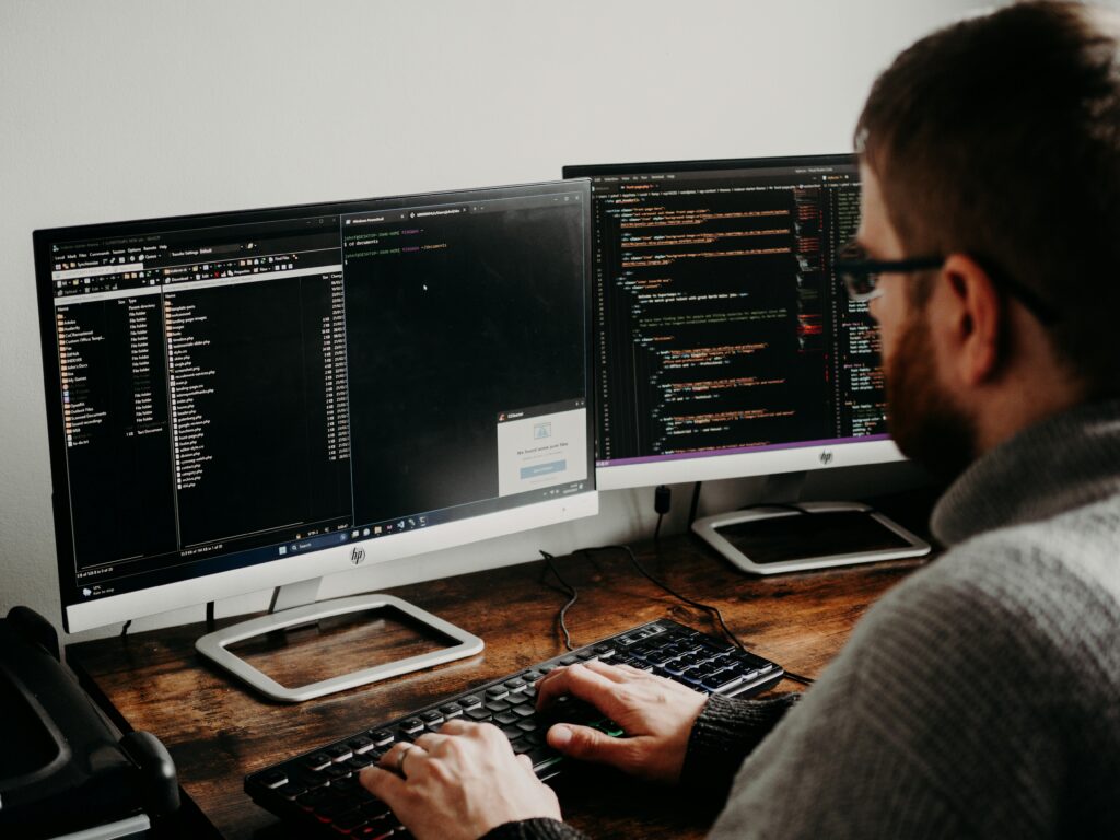 Desenvolvedor programando em duas telas com foco em back-end, escrevendo códigos e gerenciando arquivos de servidor para aplicações web.