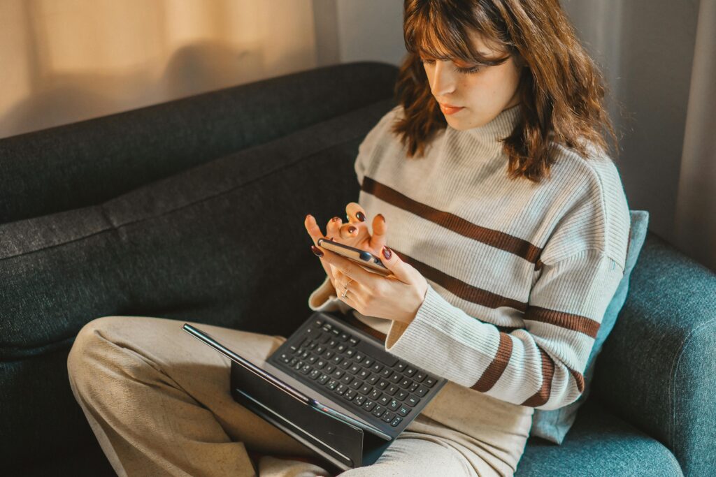 Mulher sentada em sofá utilizando celular e notebook ao mesmo tempo, representando um cenário de teste de usabilidade e análise da experiência do usuário.