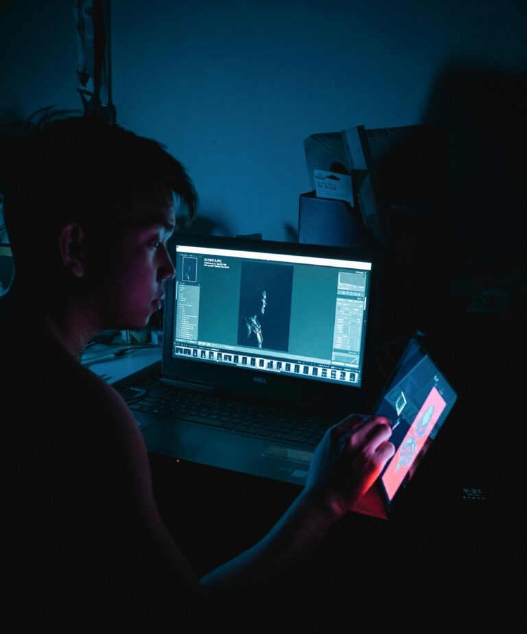 Pessoa trabalhando em ambiente escuro, usando um notebook com software de edição de imagens e, simultaneamente, um tablet para visualização de gráficos ou fluxos digitais, simbolizando a integração entre ferramentas na digitalização de processos.