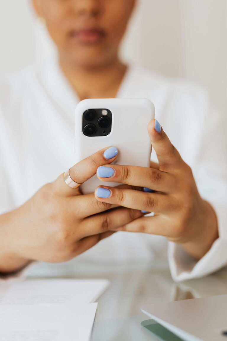 Pessoa com unhas pintadas de azul claro segura um smartphone moderno com capa branca, olhando para a tela em um ambiente interno. A imagem foca nas mãos e no aparelho, com fundo desfocado e papéis sobre a mesa.