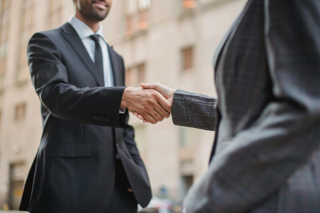Dois profissionais em trajes formais apertando as mãos em ambiente urbano, simbolizando relacionamento duradouro e confiança entre empresa e cliente.