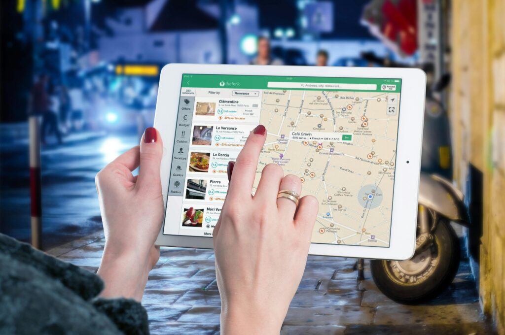 Pessoa segura um tablet e interage com um mapa digital na tela, mostrando restaurantes, locais e avaliações na interface do aplicativo TheFork, semelhante ao estilo do Google Maps, com foco em localização de estabelecimentos em uma área urbana à noite.