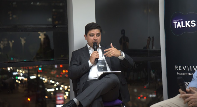 Renato Mattos mediando painel no Executive Talks, com microfone em mãos e bloco de anotações, diante de uma janela com vista noturna da cidade.