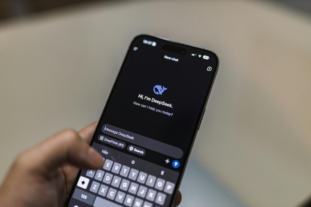 Pessoa segura um smartphone exibindo a interface do chatbot DeepSeek em modo escuro, com a mensagem de boas-vindas “Hi, I’m DeepSeek. How can I help you today?” e o campo de digitação aberto, pronto para iniciar a conversa.