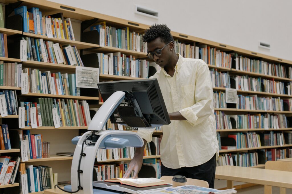 Pessoa digitalizando um livro em uma biblioteca, utilizando equipamento moderno com tela e scanner, representando o processo de digitalização de acervos físicos.