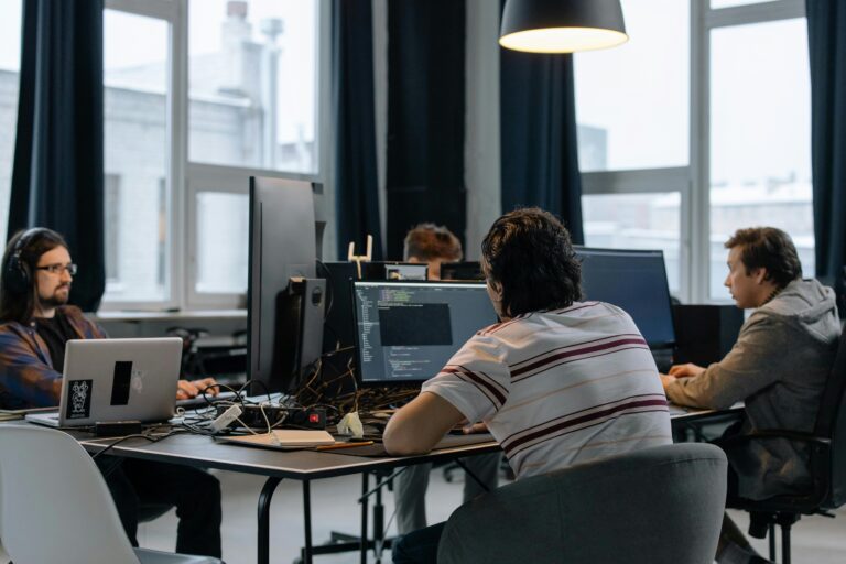 Profissionais de tecnologia sentados em frente a monitores, programando e analisando sistemas em um escritório moderno, com ênfase em processos ligados ao DNS (Domain Name System).