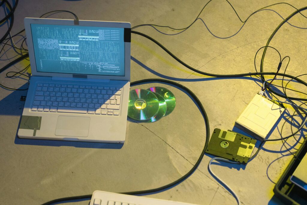 Computador portátil com interface de terminal, disquetes, CDs e cabos, representando métodos antigos e atuais de backup e armazenamento de dados.