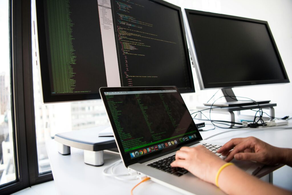Pessoa digitando em um notebook com códigos de programação abertos na tela, ao lado de dois monitores externos — um exibindo código-fonte e outro desligado, em um ambiente de escritório moderno com janelas amplas.