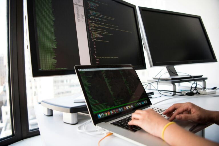 Pessoa digitando em um notebook com códigos de programação abertos na tela, ao lado de dois monitores externos — um exibindo código-fonte e outro desligado, em um ambiente de escritório moderno com janelas amplas.