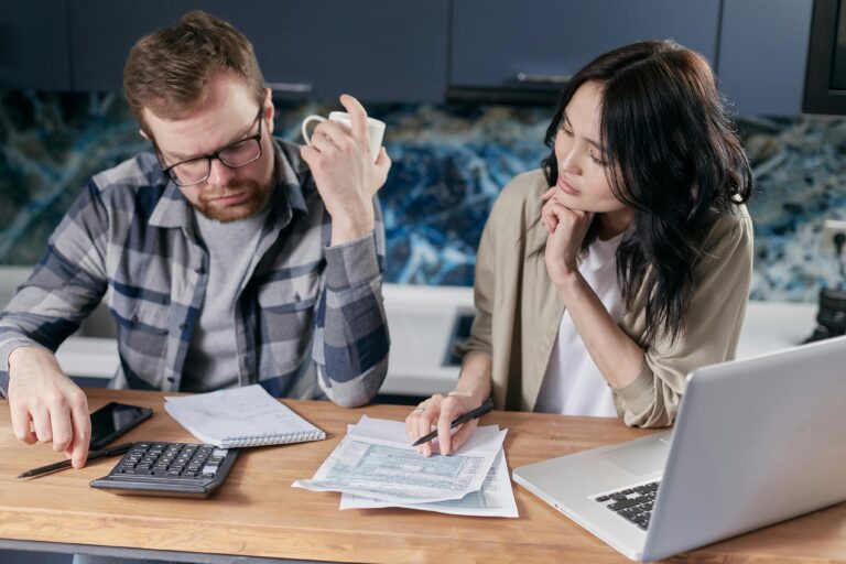 Casal analisando documentos fiscais e fazendo cálculos com notebook e calculadora, preocupado com os impactos da reforma tributária.