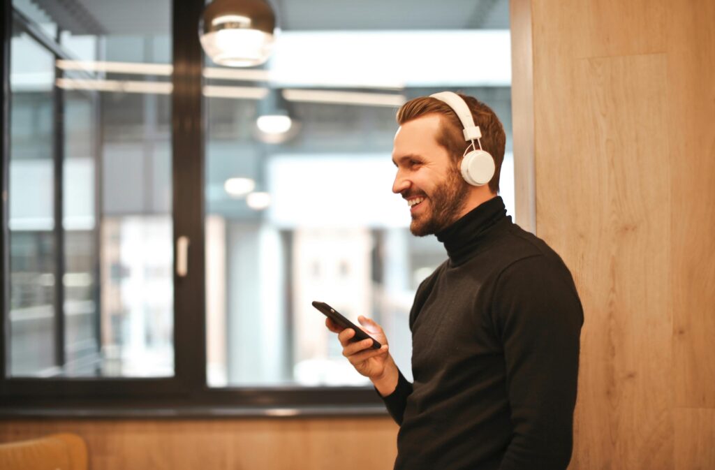 Homem sorridente usando fones de ouvido brancos conectados via Bluetooth enquanto segura um smartphone em ambiente moderno.