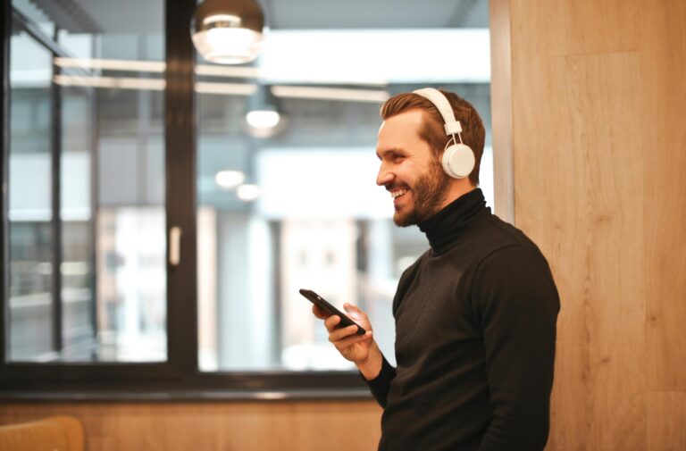 Homem sorridente usando fones de ouvido brancos conectados via Bluetooth enquanto segura um smartphone em ambiente moderno.