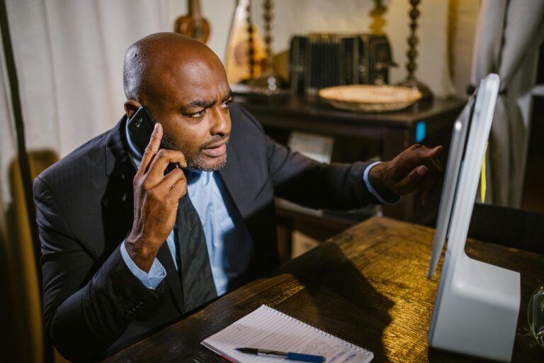 Profissional em traje formal falando ao telefone enquanto analisa algo no computador e faz anotações, representando a aplicação prática de algoritmos em decisões corporativas.