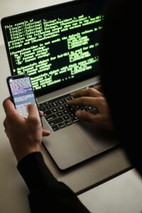 Pessoa operando um notebook com códigos verdes exibidos na tela e linhas de programação também visíveis em um smartphone, em ambiente escuro, representando atividades típicas de um hacker ou especialista em segurança cibernética.