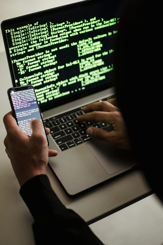 Pessoa operando um notebook com códigos verdes exibidos na tela e linhas de programação também visíveis em um smartphone, em ambiente escuro, representando atividades típicas de um hacker ou especialista em segurança cibernética.