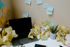 Mesa desorganizada com laptop fechado, papéis amassados e anotações adesivas coladas na parede, simbolizando uma caixa de entrada (inbox) sobrecarregada e acúmulo de tarefas pendentes.