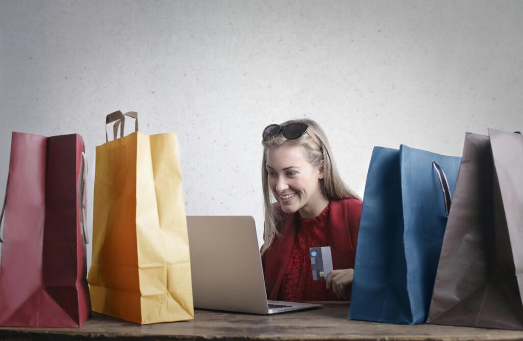 Mulher sorridente realizando compra online com cartão de crédito, cercada por sacolas de compras, representando a modernização dos pagamentos digitais com Click to Pay e Passkeys no e-commerce brasileiro.