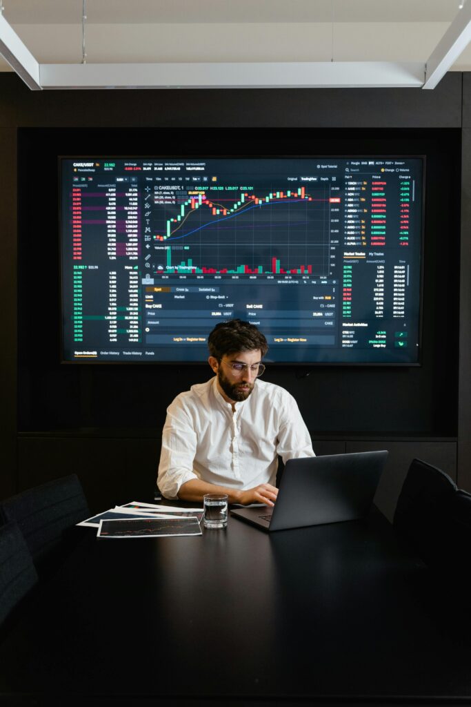 Profissional analisando gráficos financeiros em um notebook com tela grande ao fundo exibindo dados de mercado em tempo real.