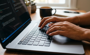 Uma foto de close-up das mãos de uma pessoa com unhas pintadas de rosa e anéis de ouro, digitando em um teclado de MacBook Pro. A tela exibe linhas de código de computador, representando o desenvolvimento e a implementação de tecnologias de Inteligência Artificial que estão impulsionando a reestruturação e a automação de empregos na Amazon.