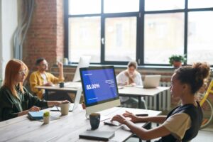 Vista de um escritório moderno com profissionais trabalhando em computadores, onde uma das telas exibe dados de desempenho em um gráfico azul, simbolizando o uso de Inteligência Artificial na análise de processos de negócios.