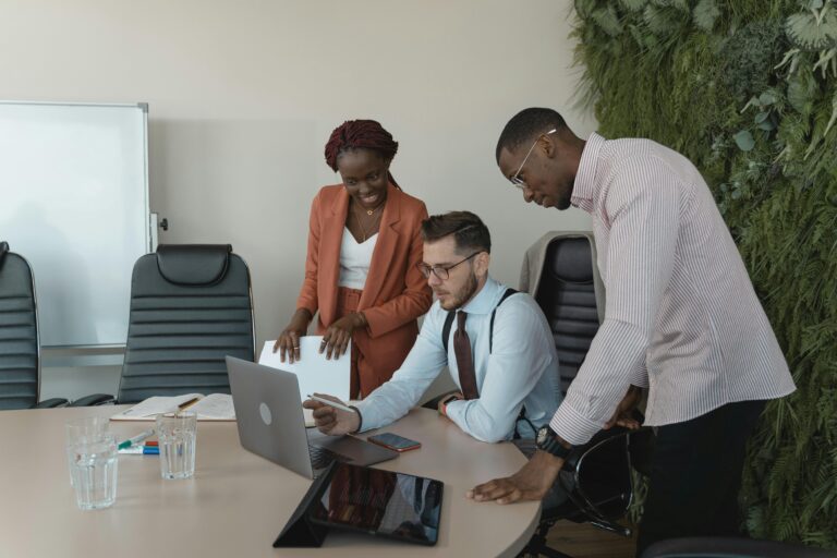 Um grupo diversificado de três profissionais de negócios colaborando ao redor de uma mesa com notebook aberto e tablet, representando a governança humana necessária para implementar sistemas de Inteligência Artificial na empresa.