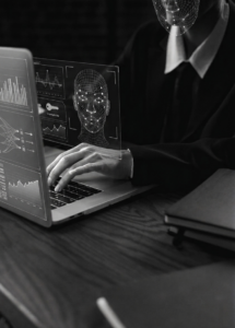Imagem em tons de cinza mostrando as mãos de uma pessoa de terno digitando em um laptop. Sobre a tela, uma interface holográfica exibe o mapeamento 3D de um rosto humano e gráficos de dados. O rosto da pessoa na imagem também está coberto por uma malha digital de rastreamento facial.