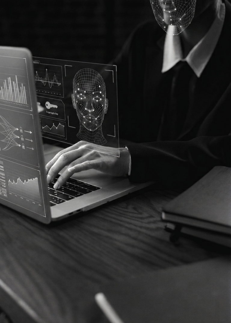 Imagem em tons de cinza mostrando as mãos de uma pessoa de terno digitando em um laptop. Sobre a tela, uma interface holográfica exibe o mapeamento 3D de um rosto humano e gráficos de dados. O rosto da pessoa na imagem também está coberto por uma malha digital de rastreamento facial.