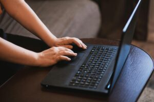 Mãos de uma pessoa digitando em um notebook preto sobre uma mesa de madeira, acessando ferramentas de inteligência artificial online.
