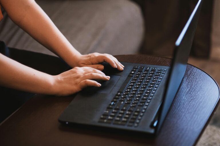 Mãos de uma pessoa digitando em um notebook preto sobre uma mesa de madeira, acessando ferramentas de inteligência artificial online.
