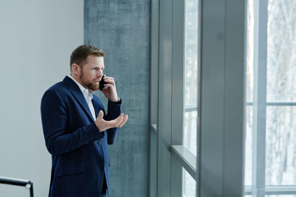 Homem de terno azul gesticulando preocupado ao telefone perto de uma janela grande, ilustrando a reação de uma vítima ao receber chamadas de golpes virtuais por clonagem de voz.